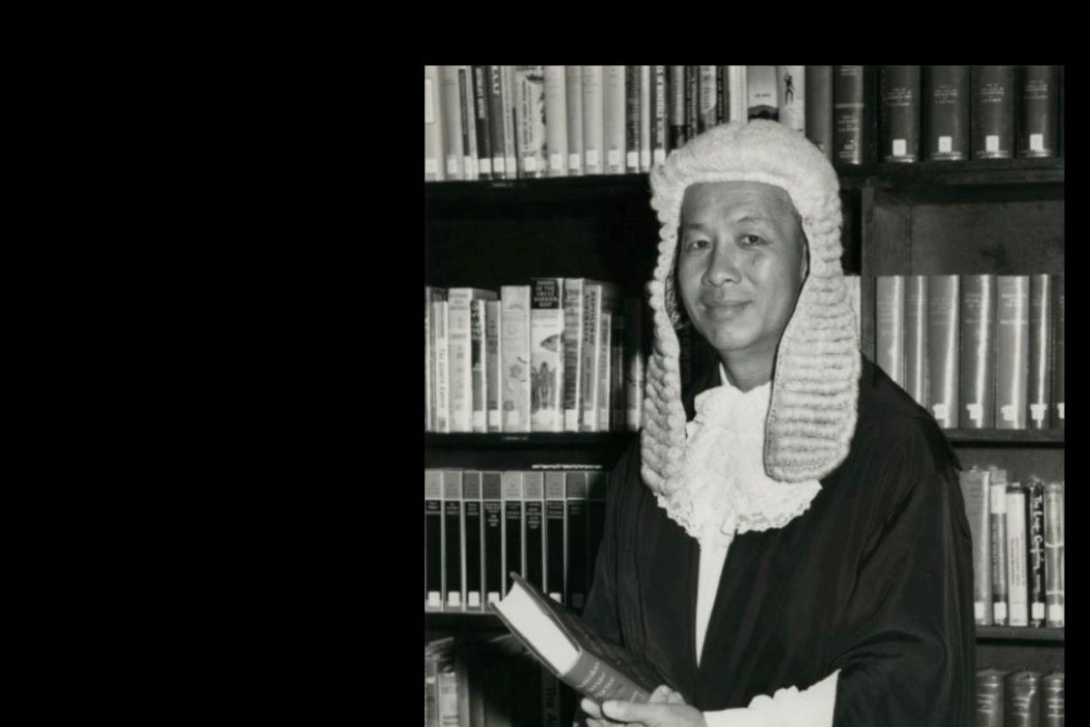 Black and white photo. Man in court wig and gown standing in front of a bookcase and holding a book.