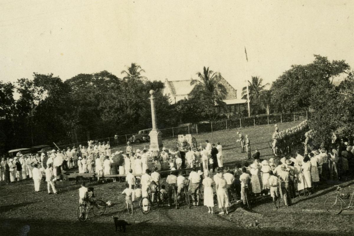 A large crowd of men and women surround the Cenotaph at centre. At far right is a line of troops, the Darwin Garrison. In the background is Government House and a flagpole with flag. In the foreground at right is a pushbike.