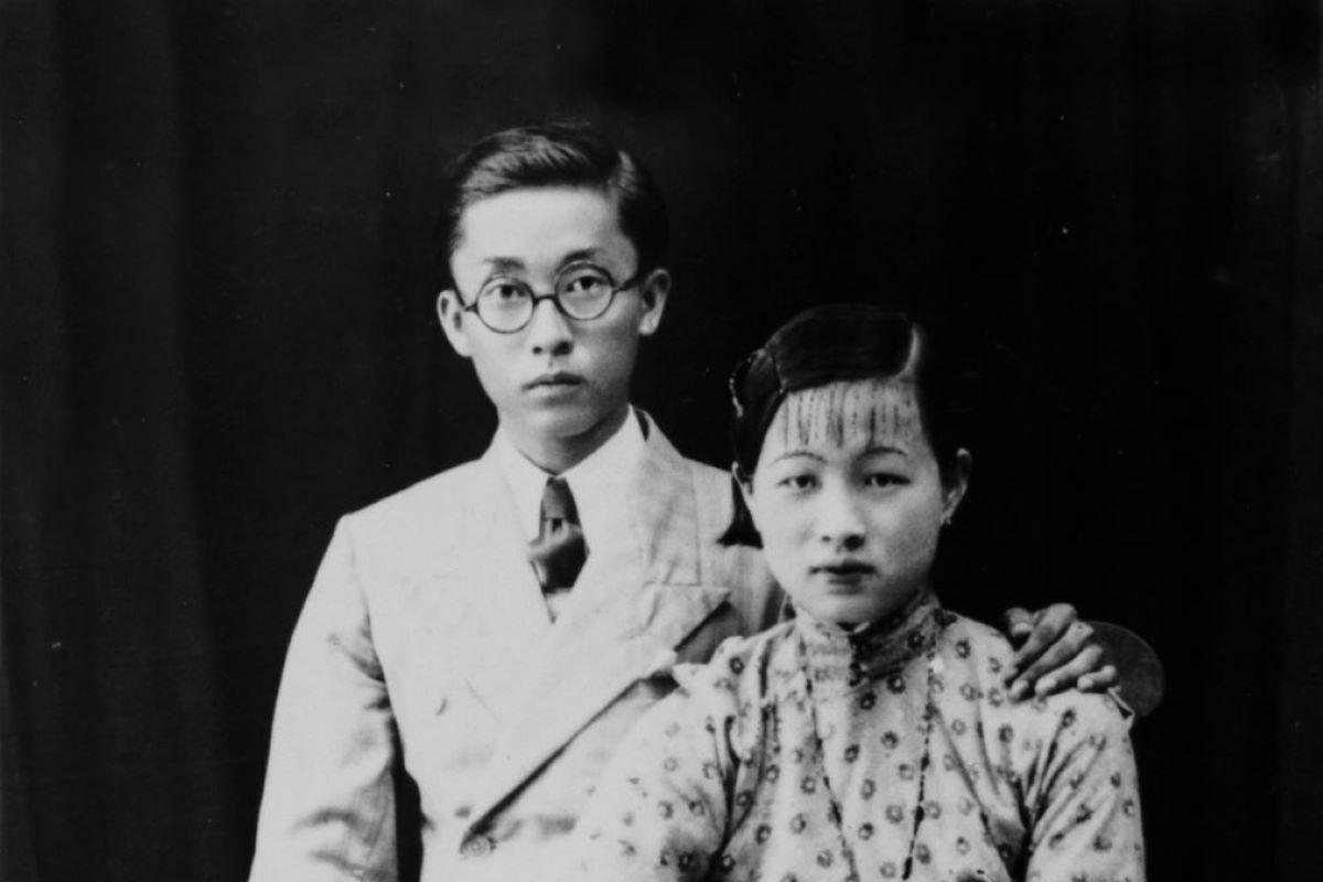 Black and white studio portrait. Man and woman are both seated, the man has this arm around the woman’s shoulder