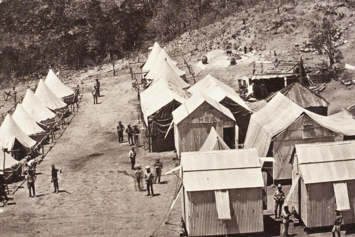 Elevated view of small corrugated iron and bark huts and large canvas tents in “street” formation with men standing outside. A large sign on one tent saying “Theatre”. 
