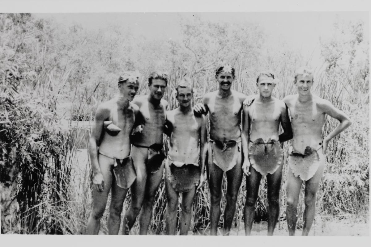 Six men standing shoulder to shoulder, wearing only lily-pad “loin cloths” and lilies in their hair.