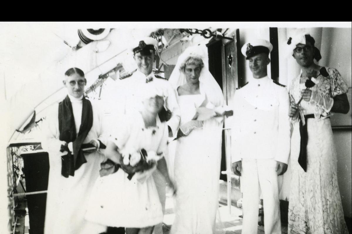 Six men on the deck of a ship dressed as a wedding party including pastor, bridesmaids and bride. 