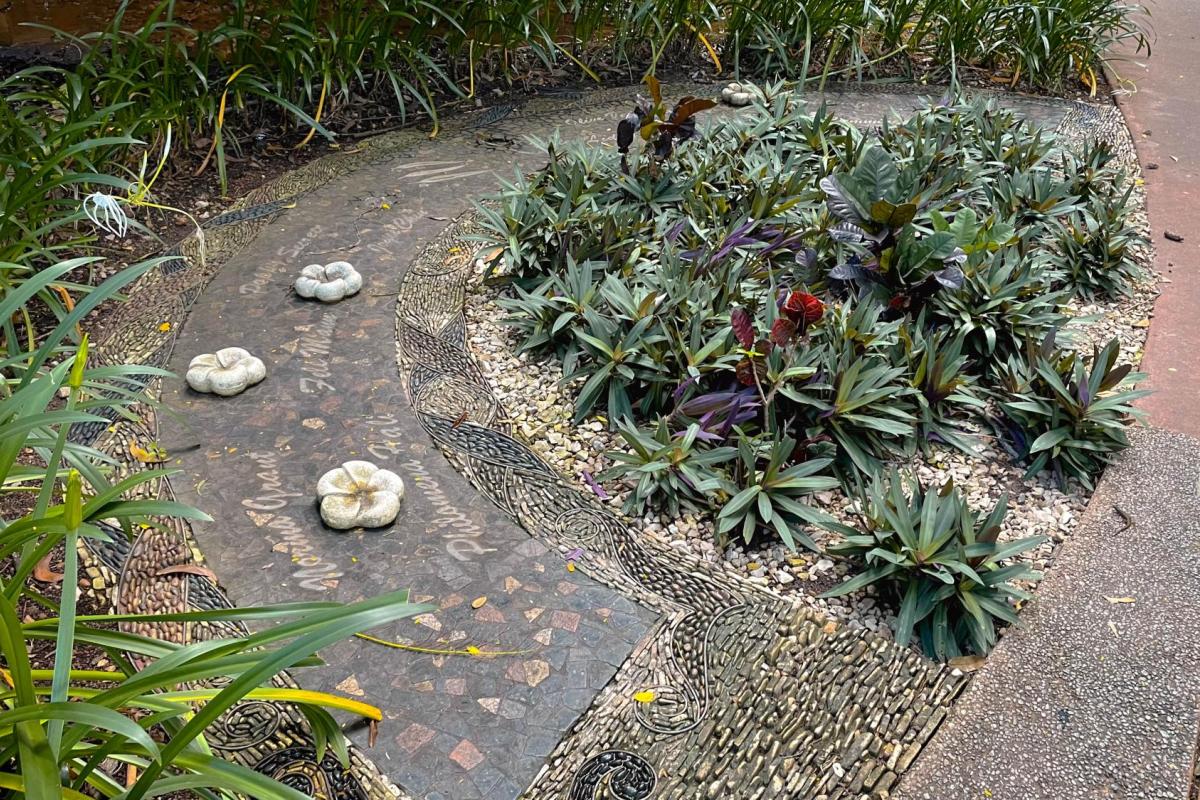 Photo of a mosaic pathway in Damoe-Ra Park. The pathway is arched with stone frangipani flowers sitting a top the mosaic. The path has the names of women who have been honoured on it in mosaic. 