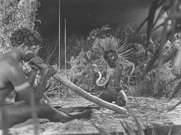 on a film set, one man playing the didgeridoo and another holding a snake