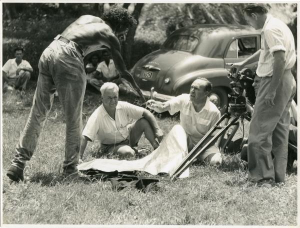 one man standing, leaning over to shake hands with a man sitting in the grass. Another man seated looking out and two other men behind a film camera