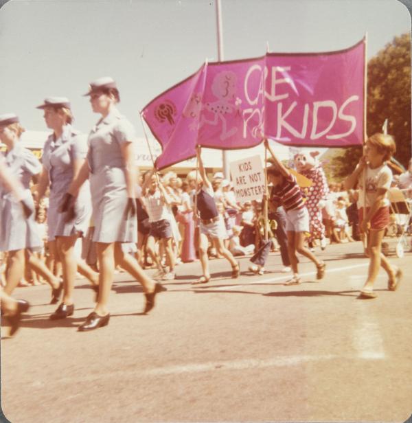 Uniformed nurses walking in a parade followed by children holding a 'care for kids' sign