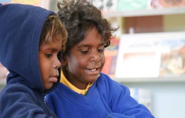 Photo taken inside a library. Two young Aboriginal boys standing at a desk reading a comic book together. Behind is a book display.