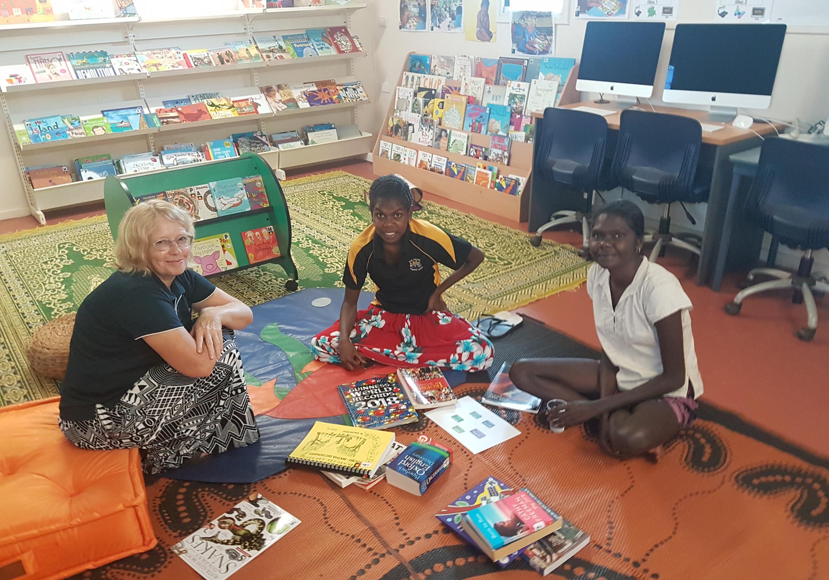 Three women sitting on the floor of a community Library space with books laid out in front of them.