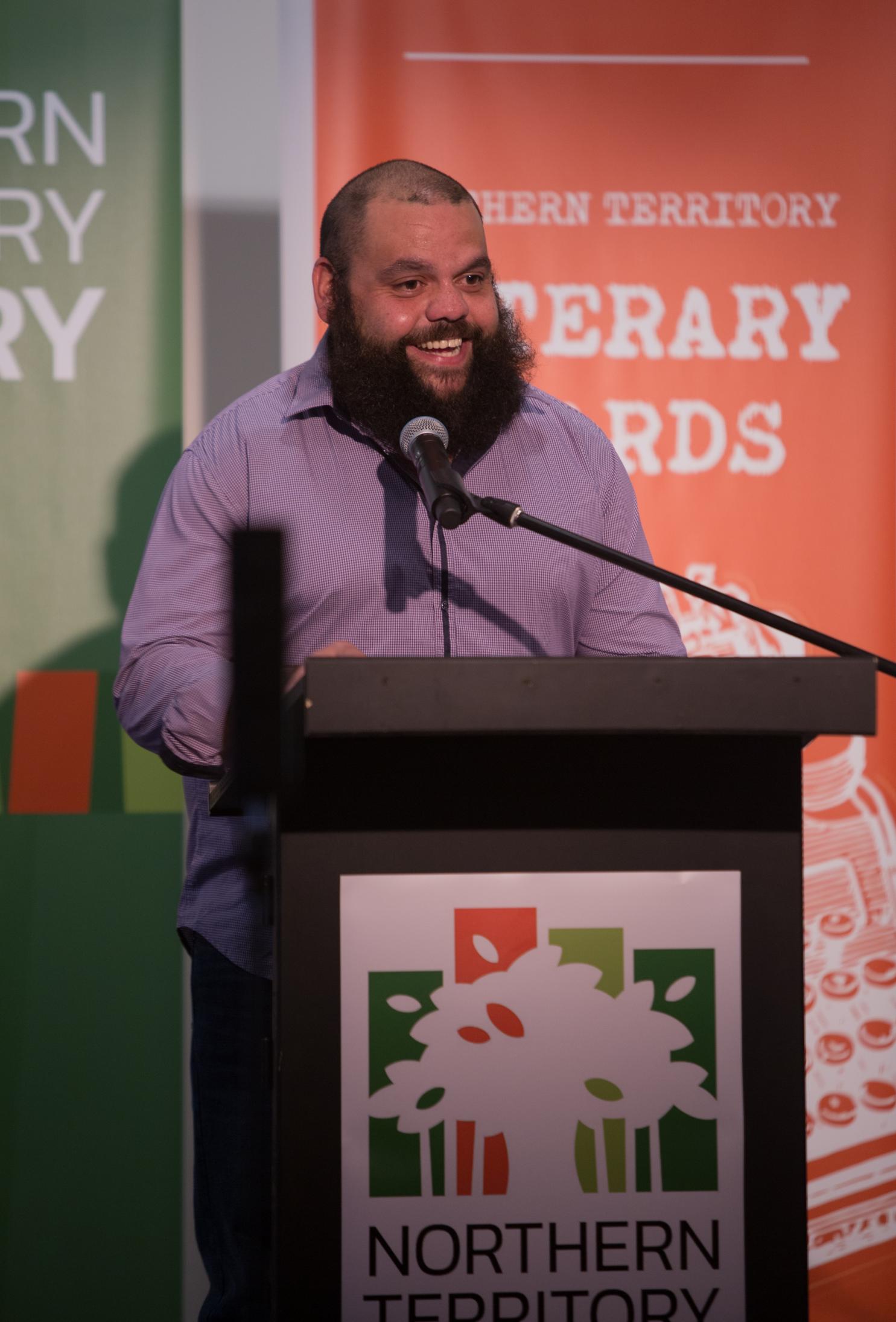 Man wearing purple shirt with a large bushy beard standing behind a lectern smiling and talking with audience. Behind him are green and orange banners. 