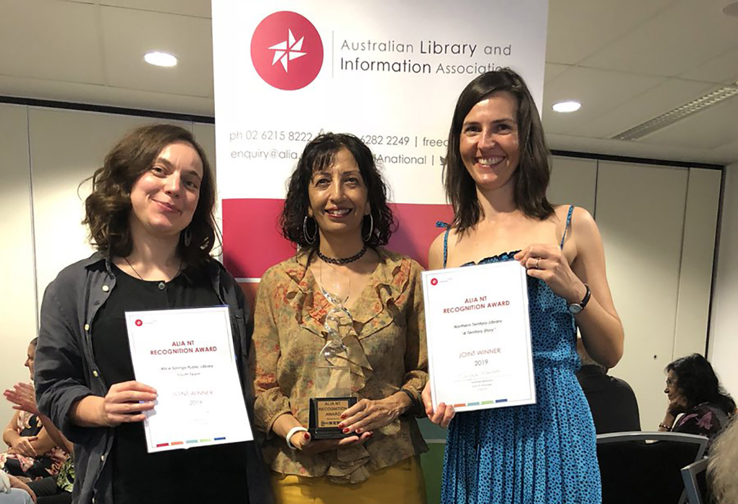 Three women standing in front of banner, holding award and certificates