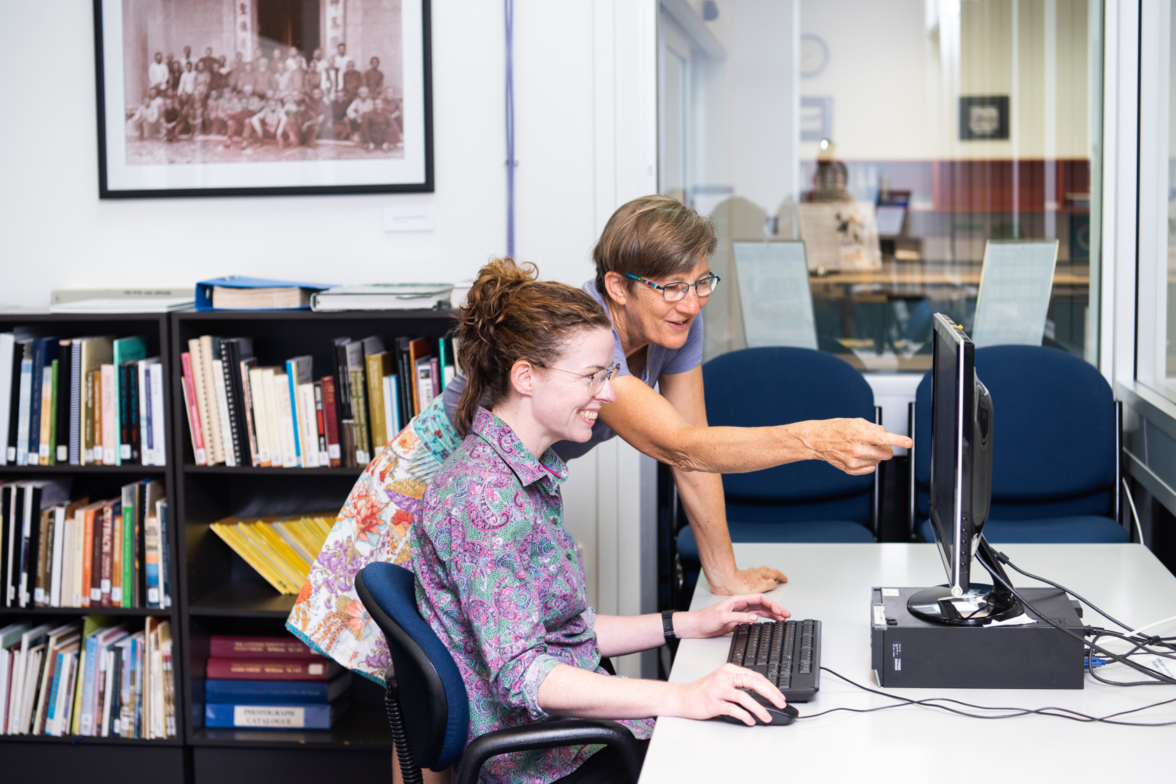 color photo of two people, one person at a pc and another providing assistance in a research setting