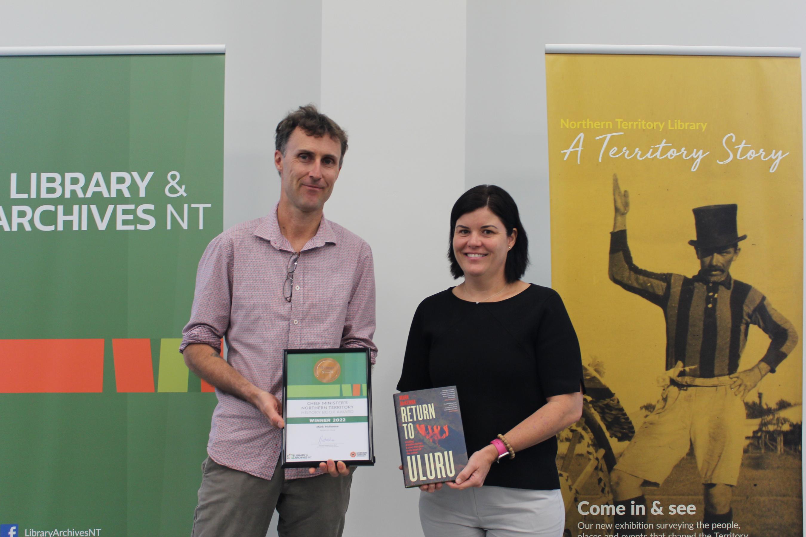 Image of tall man of causasian appearance standing next to slightly shorter woman of causacian appearance with brown hair. Man is holding framed certificate, woman is holding book 'return to uluru' Standing in between two promotional banners for LANT