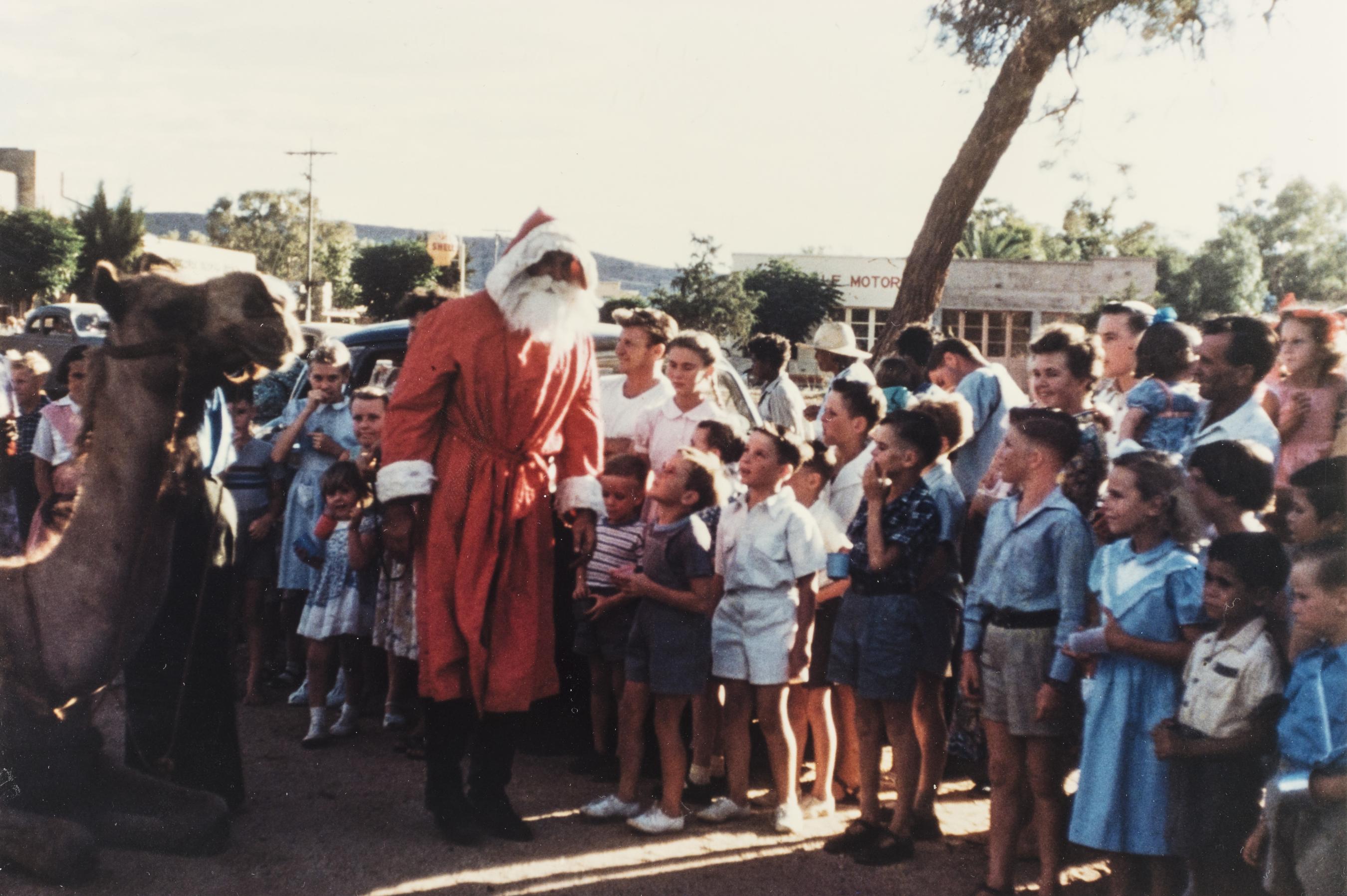 A large group of children gather around Father Christmas. A Camel is seen in the left of the frame. 
