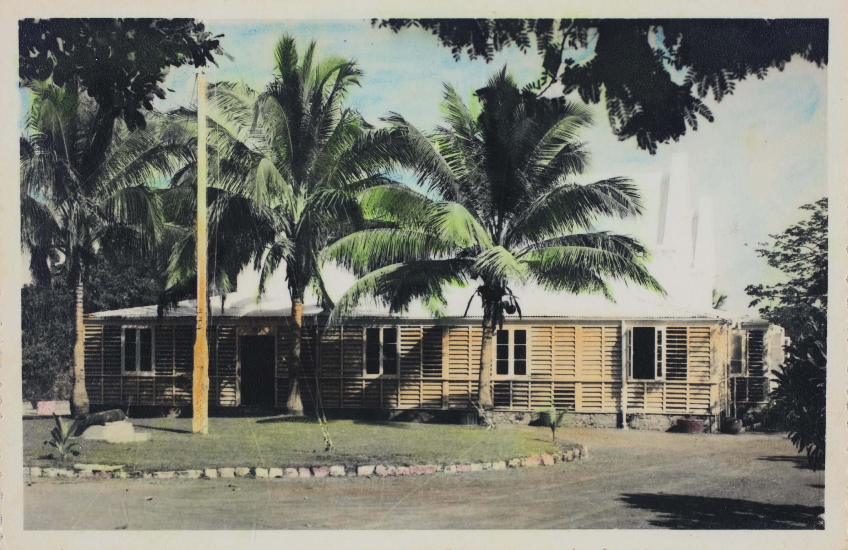 Hand coloured photograph of three large palm trees and a flagpole in front of an old tropical style building with timber louvres as the front wall. 
