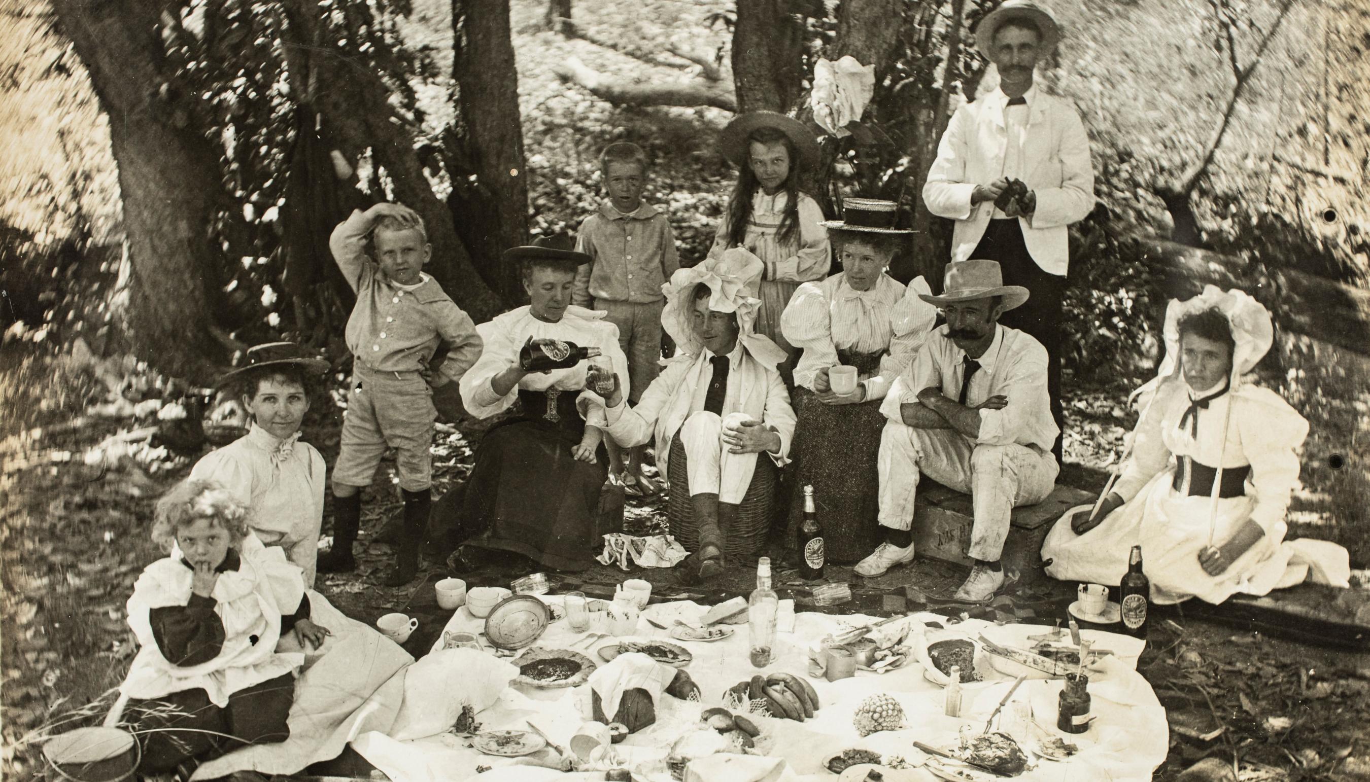 Black and white photograph of people sitting outside around a picnic blanket.