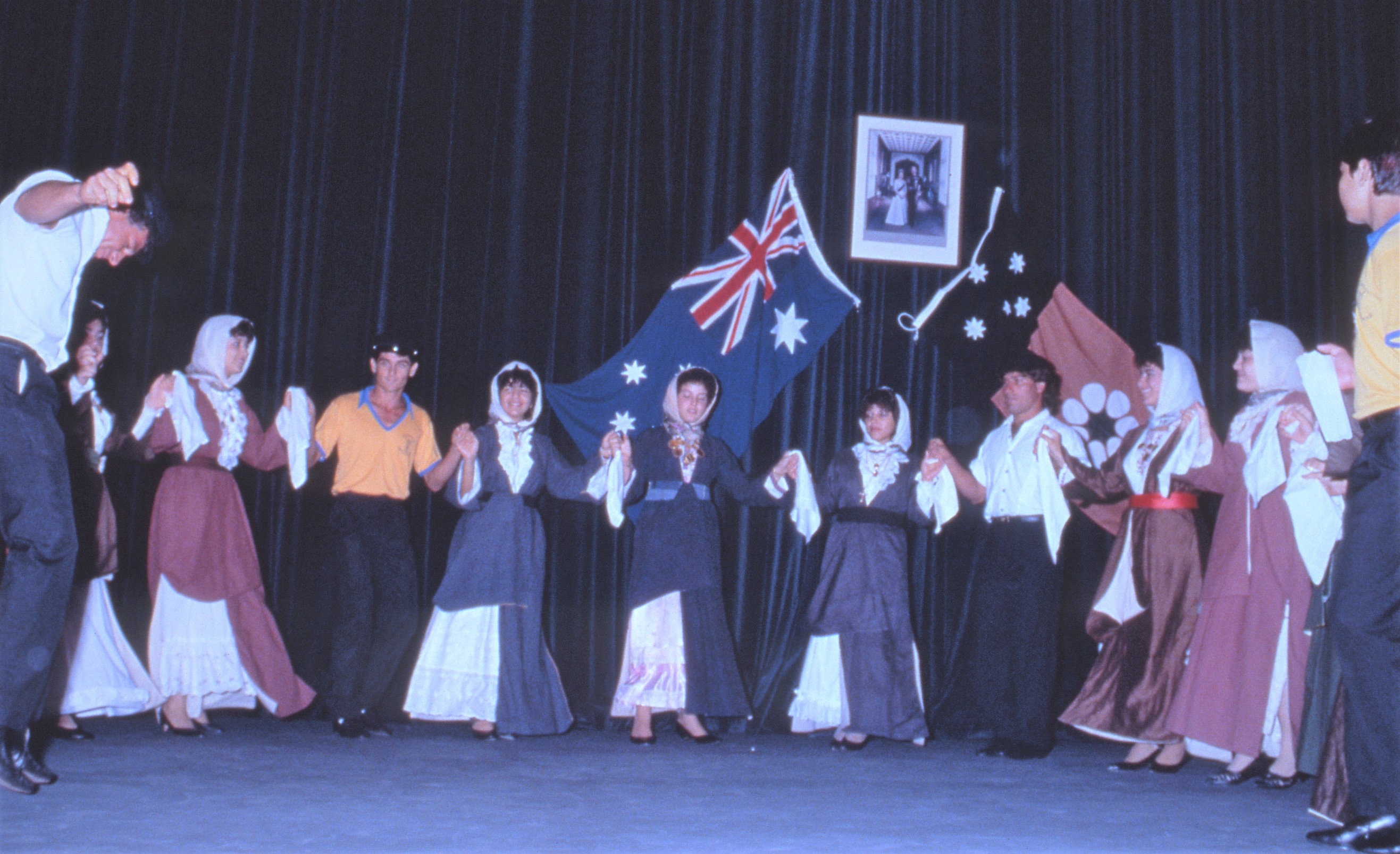 A row of eleven people in traditional Greek dress hold hands and dance in front of the Australian and Northern Territory flags. 