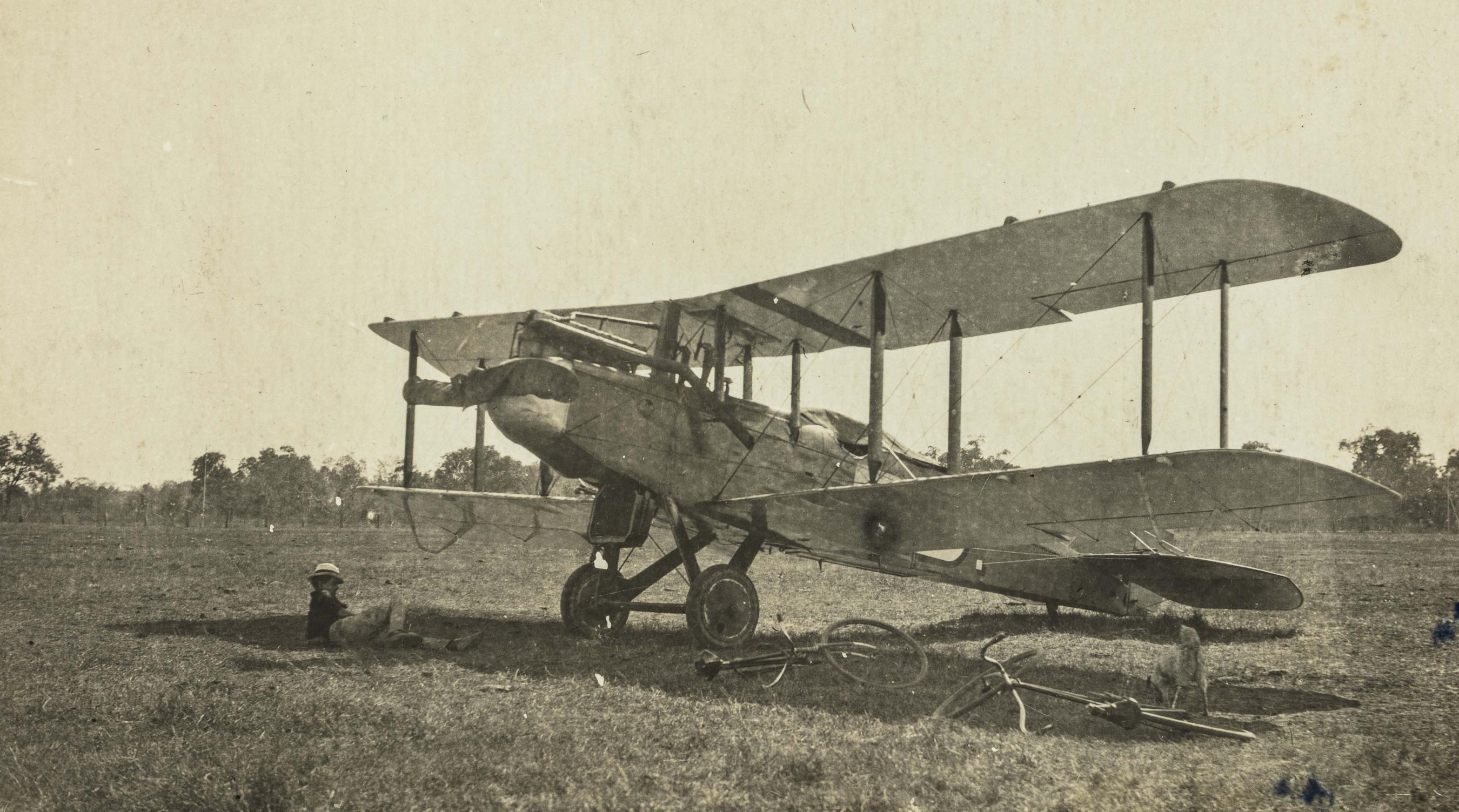 Black and white photograph of a woman laying on the grass beside a parked biplane.