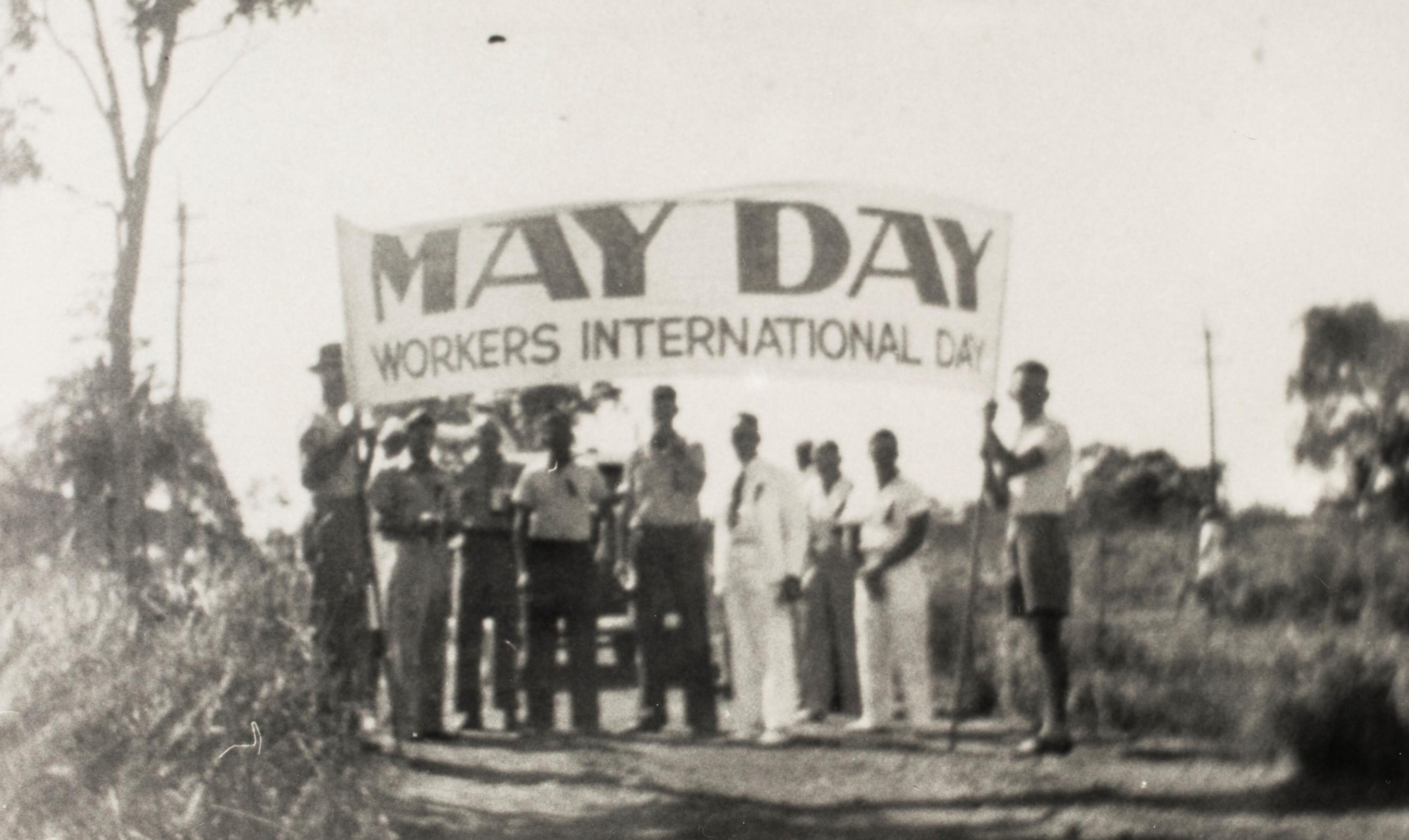 Black and white photograph of people standing beneath a May Day banner.