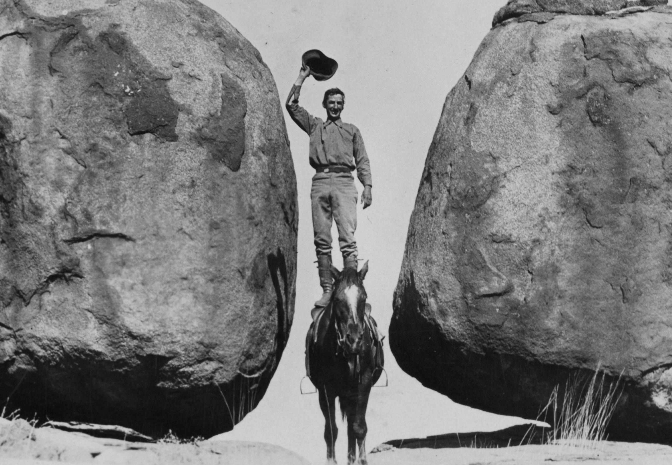 Black and white photograph of a man standing on a horse between two boulders holding a hat above his head.