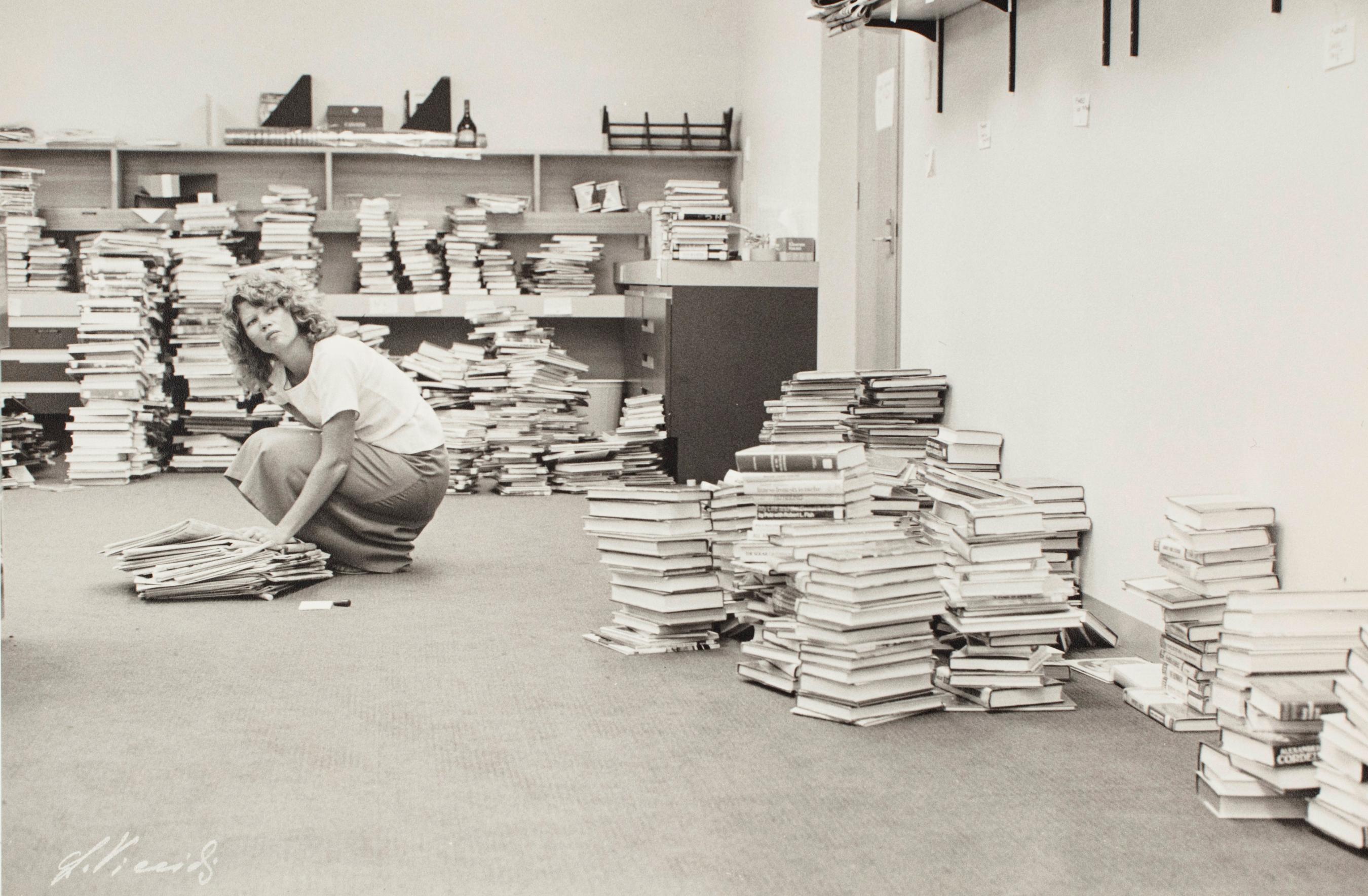 Black and white photograph of a woman crouching down among stacks of books and magazines.