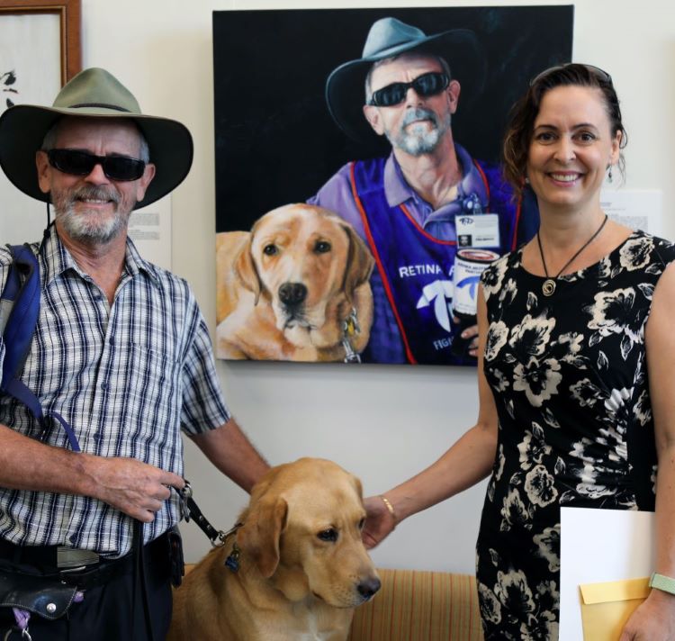 Man (Paul Patteson) with Guide Dog (Elma) and woman (Catherine Miles) standing in front of Catherine's winning artwork 'Paul and Elma'