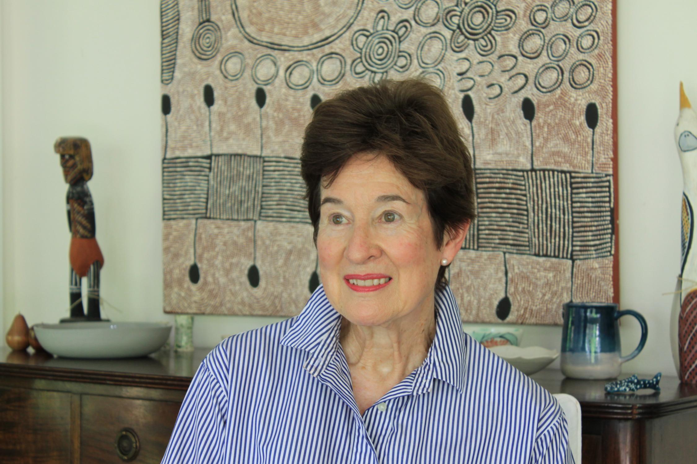 Woman looking to the left of camera wearing stripped blue and white shirt, sitting in front of an Aboriginal artwork