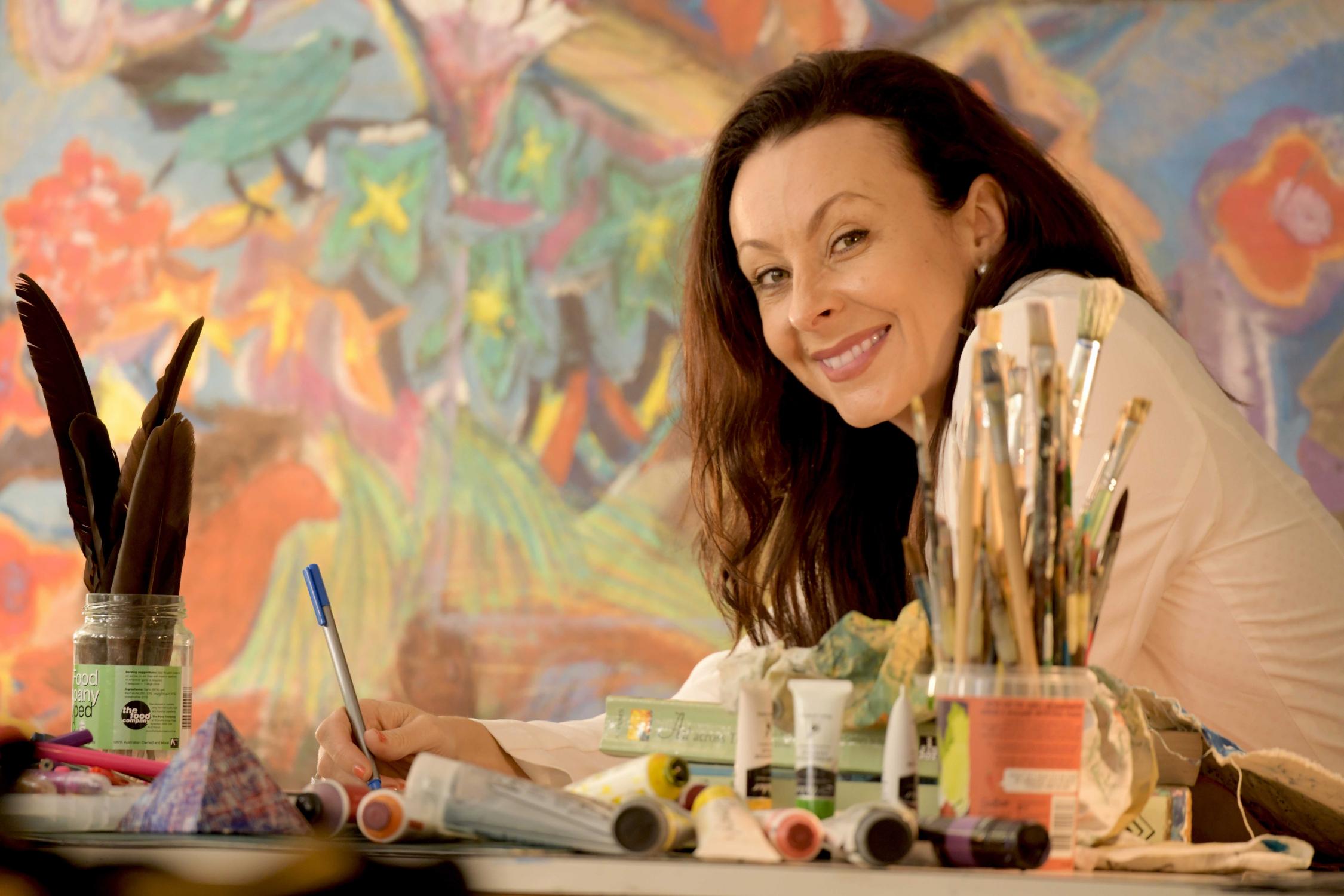 Picture of middle aged woman sitting in front of a brightly coloured painting, at a desk surrounded by paint brushes and paint. 