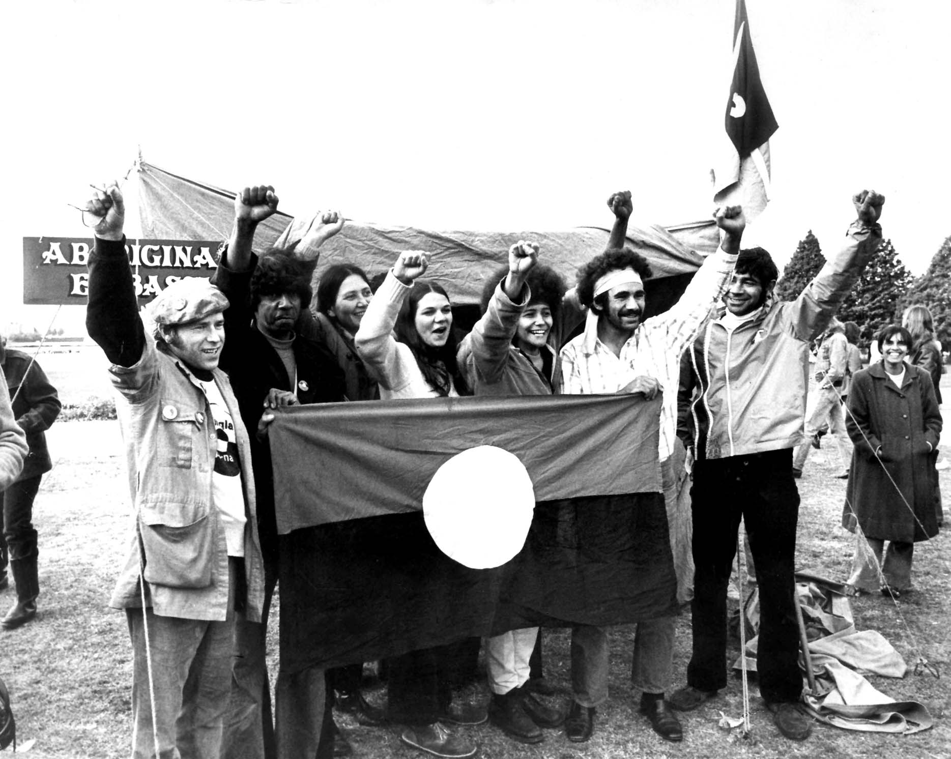 black and white image of 7 aboriginal protestors with raised fist behind the aboriginal flag - red at the top, yellow circile in the middle and black band at the bottom. 
