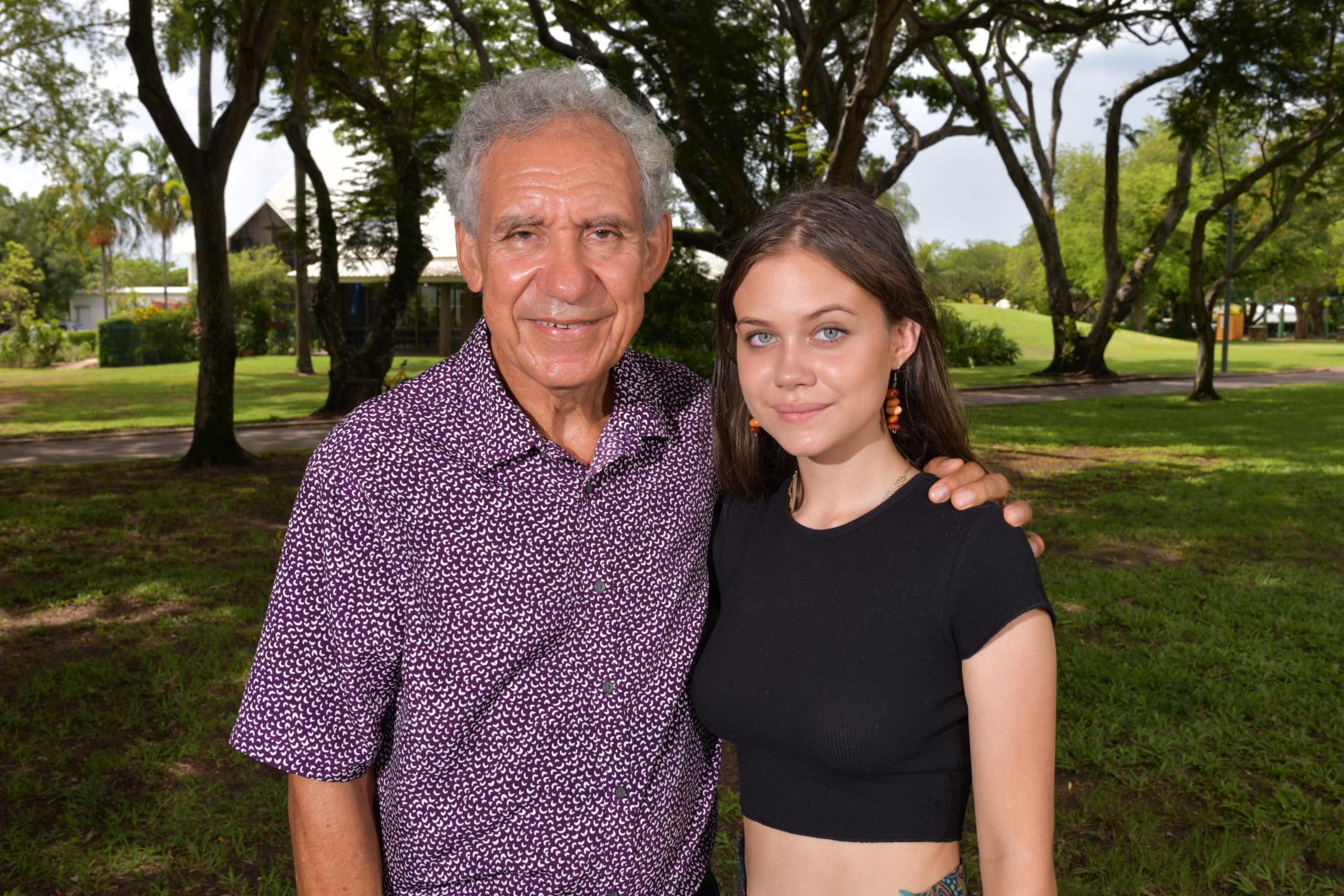 Older man with medium toned skin standing next to younger lady in a grassy field in the shade of trees