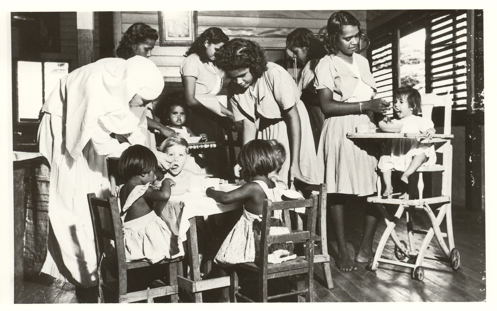 5 young adults and 1 older lady who is dressed in a nun's uniform are feedings smaller kids with dark skin who are sitting in high chairs