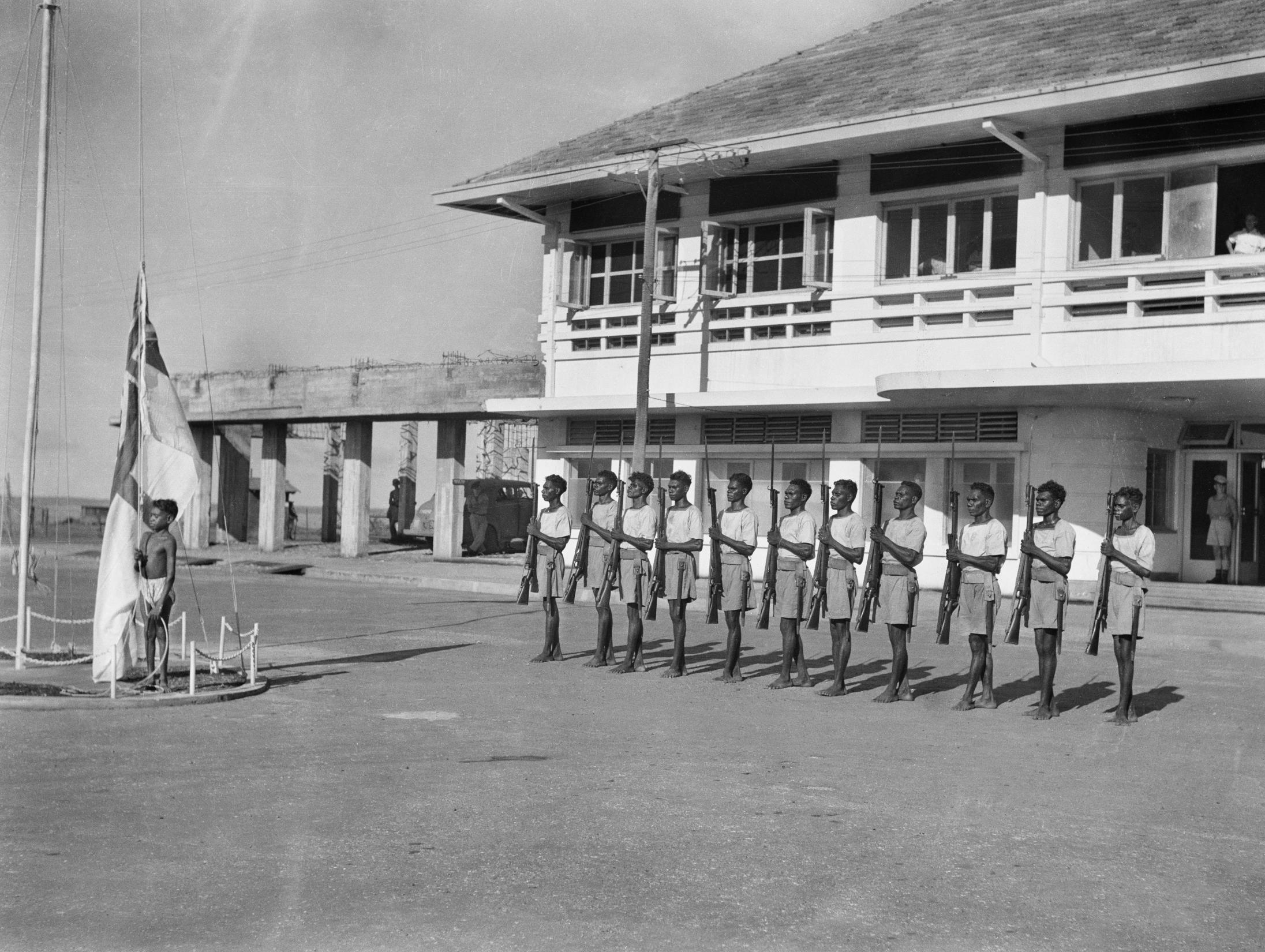 Image of black and white men standing in a line in front of a double storey building in uniform holding rifles. Boy standing just in front off to their right hand sides raising a flag. 