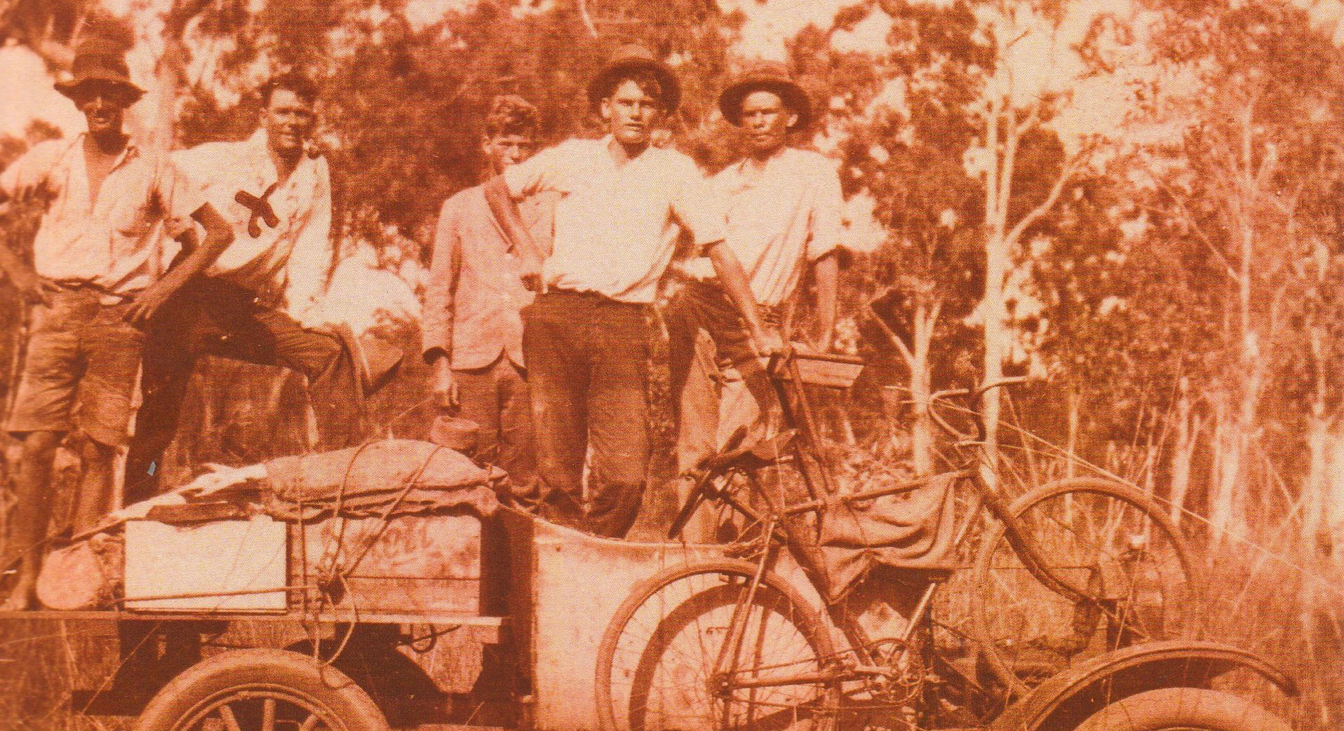 Orange toned black and white photograph of five men standing on a open top vehicle with a bicycle strapped to the front.