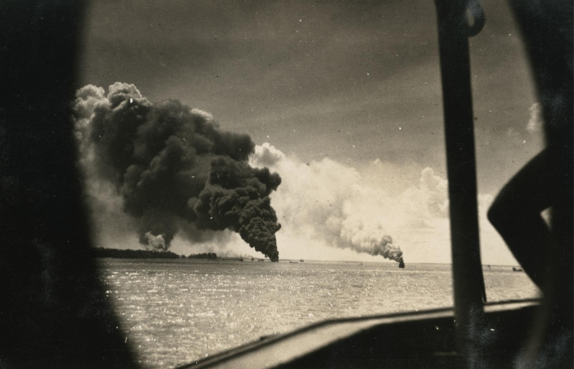 Black and white photograph of smoke plumes rising from two bombed ships on the sea.