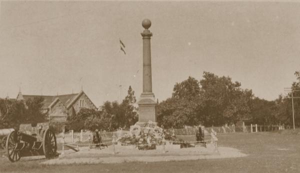 The Cenotaph stands at centre with floral wreaths at its base. In the background is Government House and two machine guns. At left is a field gun.