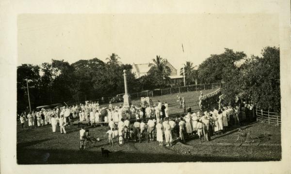 A large crowd of men and women surround the Cenotaph at centre. At far right is a line of troops, the Darwin Garrison. In the background is Government House and a flagpole with flag. In the foreground at right is a pushbike.