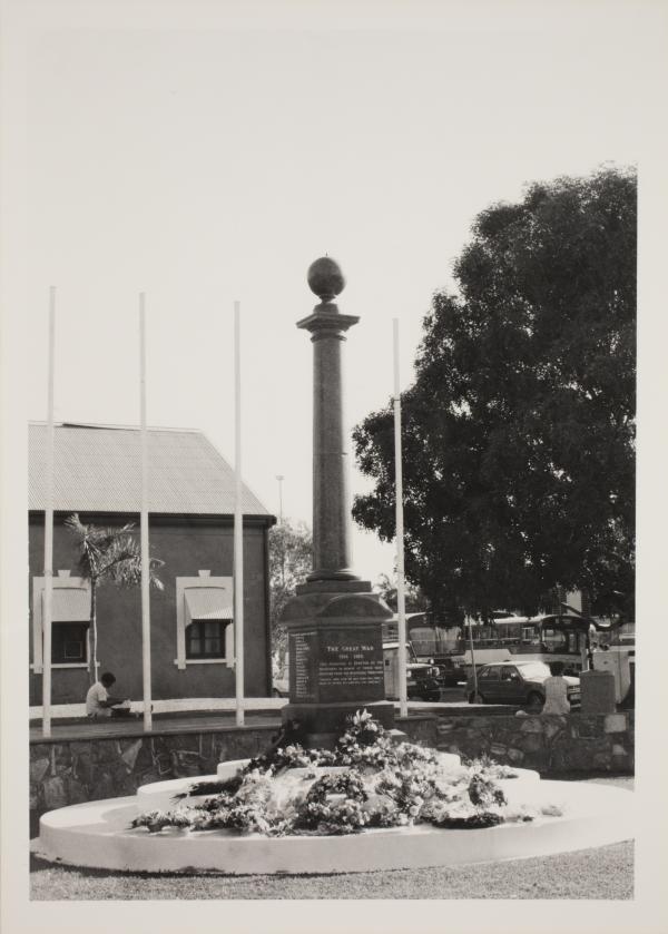The Cenotaph is at centre with floral wreaths at its base. In the background at left is a building (Browns Mart) and at right is a large tree. A bus can be seen in the far distance.