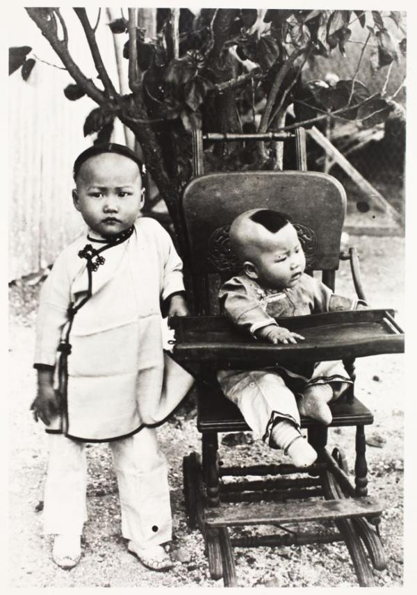 Black and white photo. A young child and a baby. The baby is seated in a wooden pushchair 