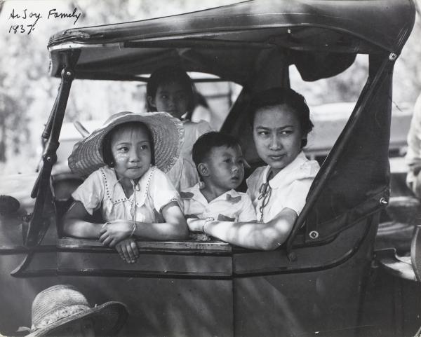 Black and white photo. Two children and a woman sitting in the front seat of an older style car. Women on the seat and two children on leaning on the dashboard. All looking out the passenger side door. 