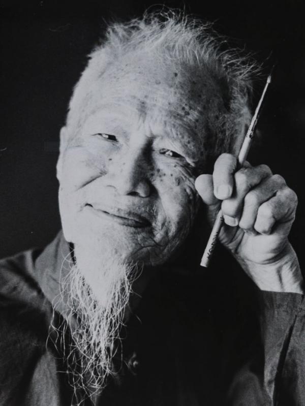 Black and white photo. An elderly man is smiling. His hand, which is held to his face grips a paintbrush between his index and forefinger 