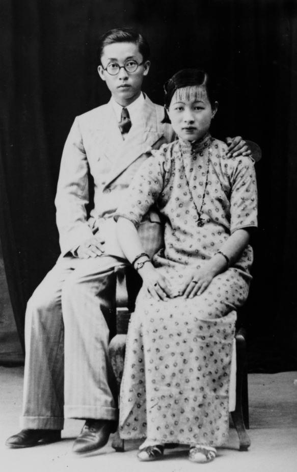 Black and white studio portrait. Man and woman are both seated, the man has this arm around the woman’s shoulder
