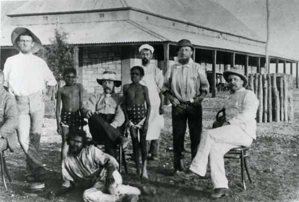Depicts a group of Aboriginal and non-Aboriginal men and boys in front of a stone building.