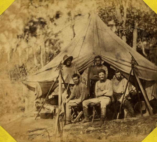 Depicts six surveyors at rest in front of an open tent with two theodolites at left and right.