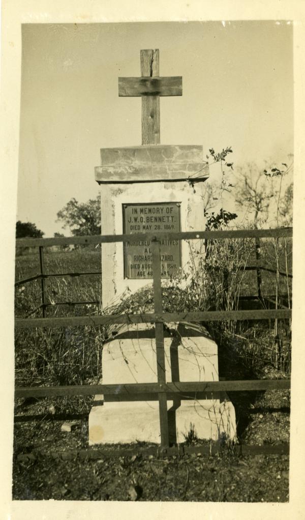 Depicts John Bennett’s grave behind a wooden fence. The grave is a high stone pedestal with a wooden cross on top and a bronze memorial plaque at front.