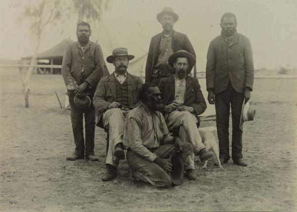 Depicts three Aboriginal men and three non-Aboriginal men posing for the camera in front of their camp.