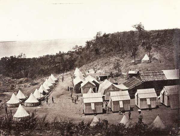 Elevated view of small corrugated iron and bark huts and large canvas tents in “street” formation with men standing outside. A large sign on one tent saying “Theatre”.