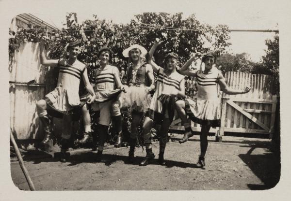 Five men in a “revue” pose, wearing short dresses, corsets, hats, make-up and boots.