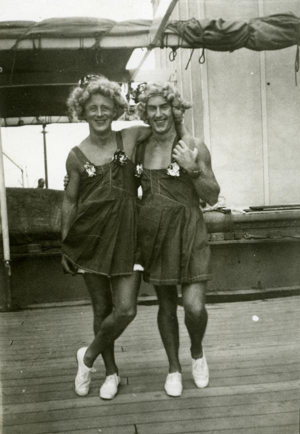 Two laughing men wearing identical short dresses, make-up and wigs on the deck of ship. One has his arm around the other’s shoulders. 
