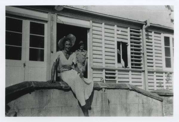 Smiling man wearing summer dress and hat sits in front of damaged building with a smiling soldier in doorway.
