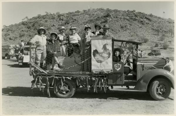 Seven masked people in women’s clothing standing on the back of a truck with a sign reading “Ye Olde Hens”.