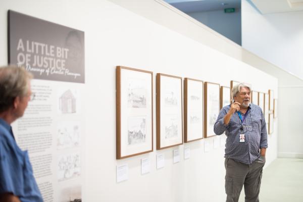 Man speaking in front of framed drawings hung on a wall. 
